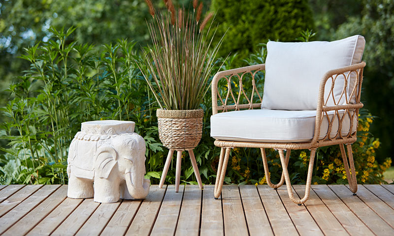 Uteplass med flettet kurvstol med hvite puter, hvit elefantkrakk og plante i kurv