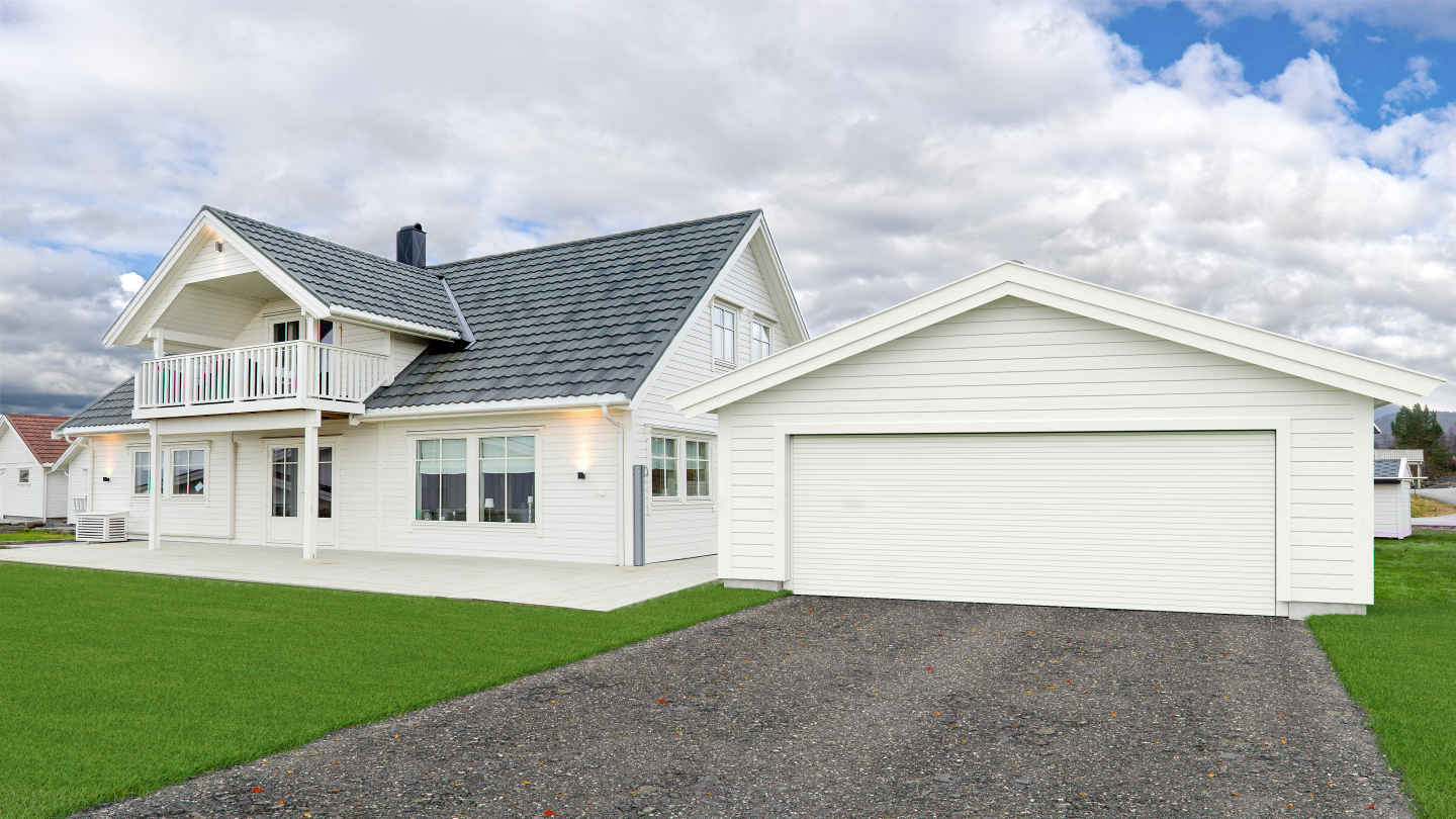 Hvit trevilla med grått tak, dobbelgarasje, grønn plen og skyet himmel.