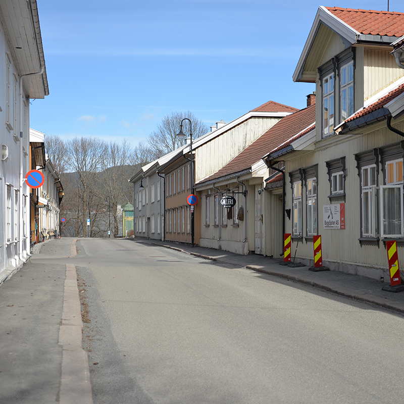 Stille asfaltert gate med lysegule trehus, røde tak, “Galleri”-skilt og klar blå himmel