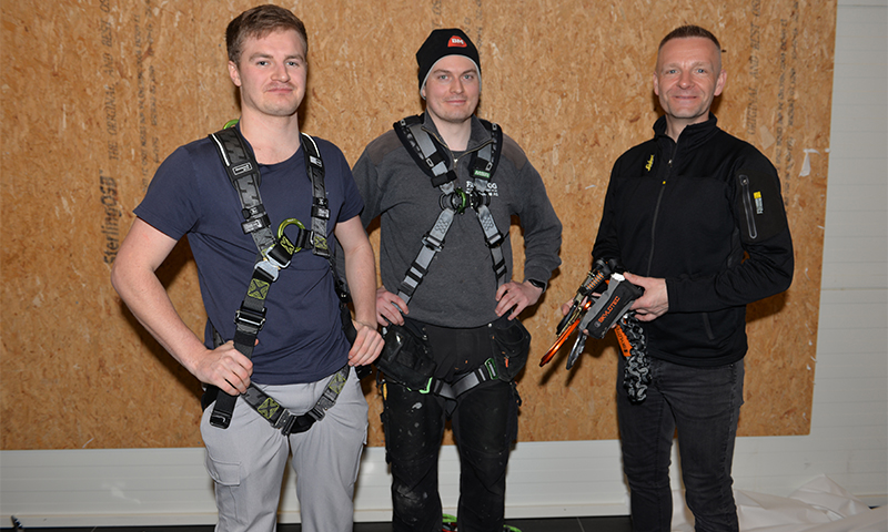 Tre menn i mørke arbeidsklær og verneseler med verktøy foran trepanel.