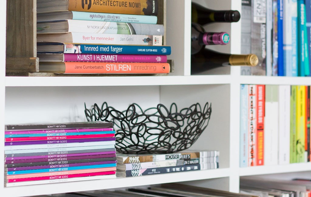 Hvit bokhylle med fargerike interiørbøker, stabler av magasiner, svart nettingbolle og vinflasker.
