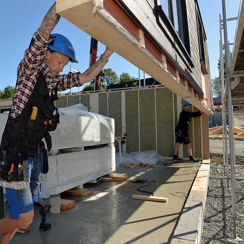 To arbeidere løfter veggmodul mot mørk trefasade på byggeplass under klar blå himmel.