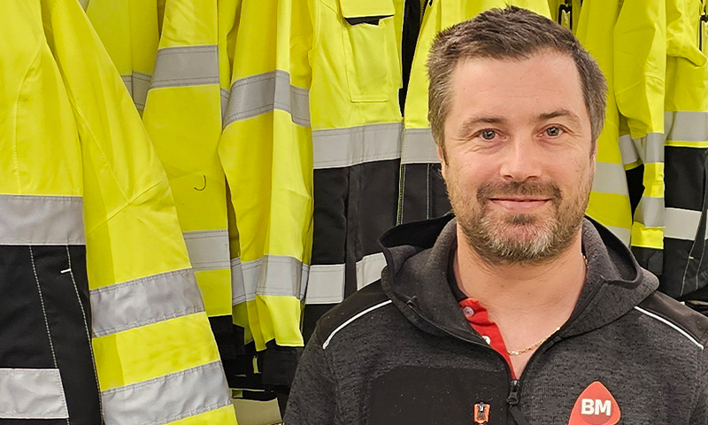 Mann i mørk jakke med BM-logo foran gule refleksjakker på stativ.