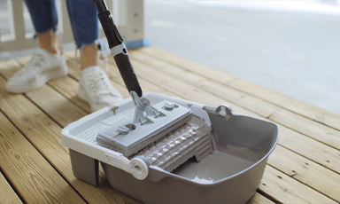 Grå roterende mopp i bøtte på lyst tregulv, jeanskledd person med hvite sneakers bak