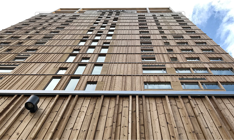 Froskeperspektiv av høyt trebygg med lyse, vertikale plankefasader og smale vinduer mot blå himmel