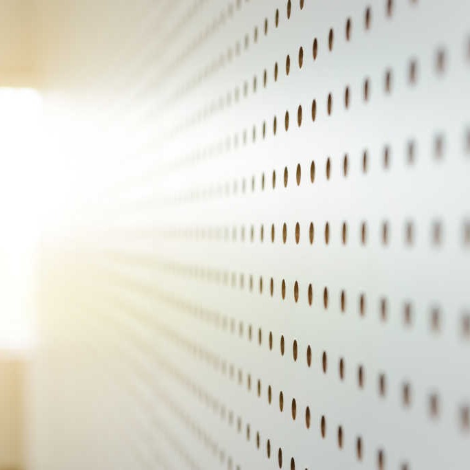 Hvitt perforert panel med rader av sirkulære hull i perspektiv, sterkt sollys fra venstre.
