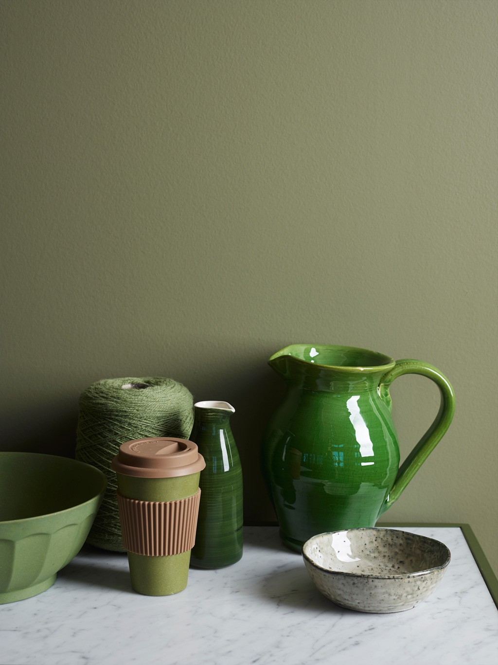 Stilleben på marmor med grønn mugge, bolle, takeaway-kopp, garnnøste mot olivengrønn vegg