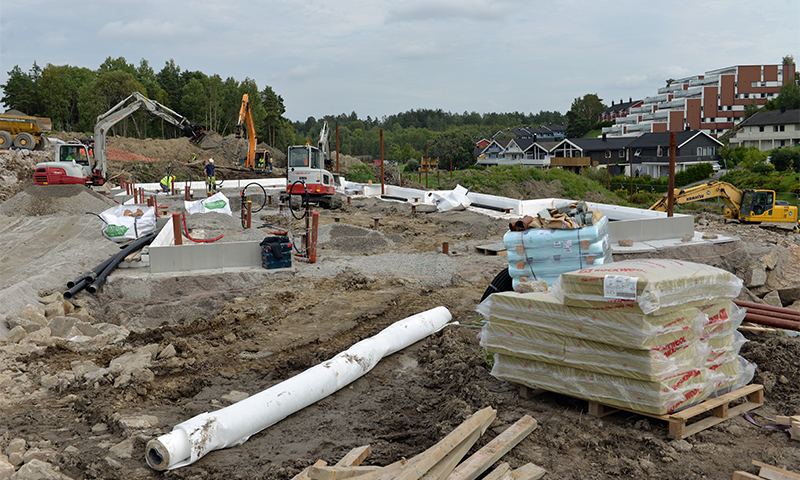 Grå byggeplass med gravemaskiner, hvite isolasjonsplater og boligblokker i bakgrunnen.