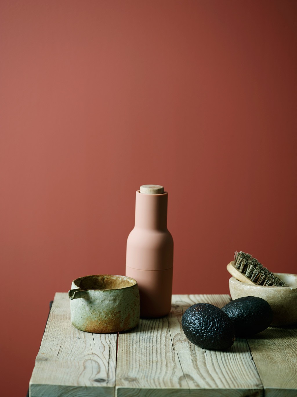 Treplate med keramisk mugge, rosa krydderkvern, to avokadoer og kost mot rødt bakteppe.