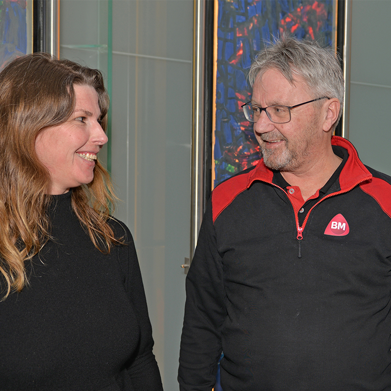 To personer, kvinne i svart genser og mann i sort-rød BM-jakke foran fargerikt glassmaleri