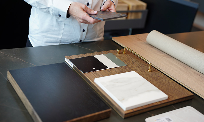 Hender holder grå flis over utvalg med svart treplanke, hvit marmorklinker, fargeprøver og beige stoffrull.