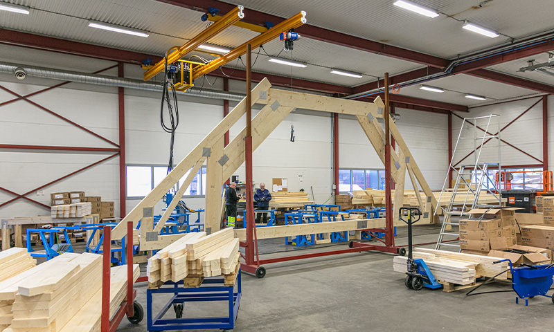 Lyst verksted med trekonstruksjon i lys furu på røde/blå stativer under gul traverskran.