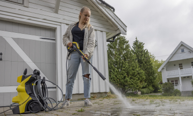 Kvinne bruker gul høytrykksvasker på grå belegningsstein foran hvitt hus og grønne busker.