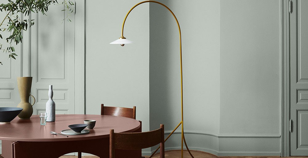 Rundt støvet rosa spisebord med keramikk, trestoler og gult buet gulvlampe mot lysegrønn vegg