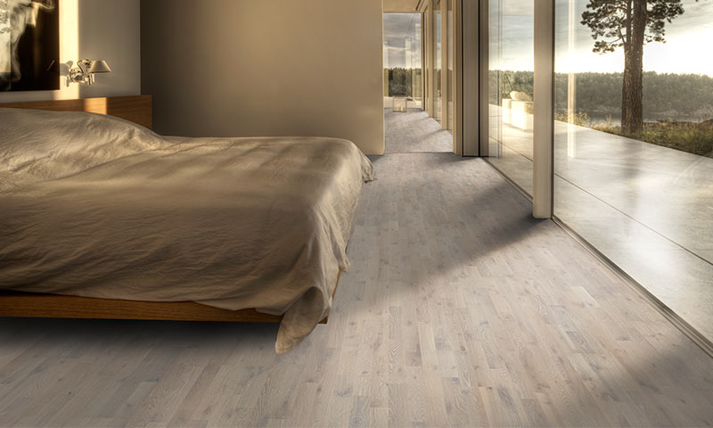 Lyst soverom med tremøbler, beige seng og store glassvinduer mot skog og innsjø.