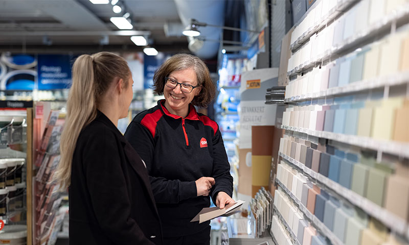 Ansatt i sort/rød uniform viser pastellfargede malingprøver til kunde i butikk.