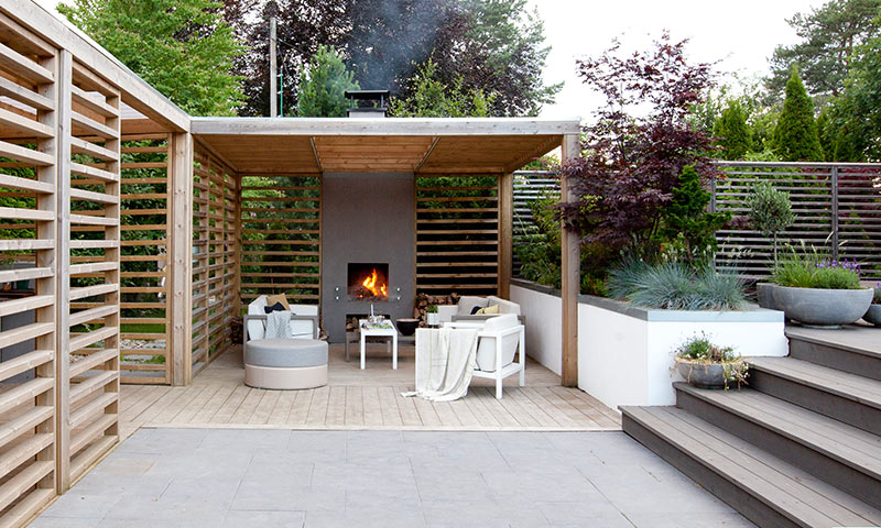 Moderne ute-lounge under lyst tretak, med grå peis i bruk, hvite møbler og grønn hage.
