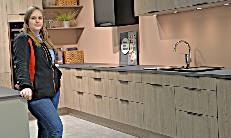 Kvinne ved moderne kjøkken med lyse tremøbler, svart benkeplate, vask og åpne hyller.