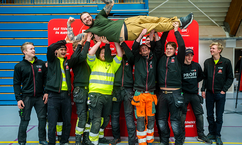Åtte håndverkere i arbeidsklær og high-vis løfter en kollega over hodet foran rødt banner.