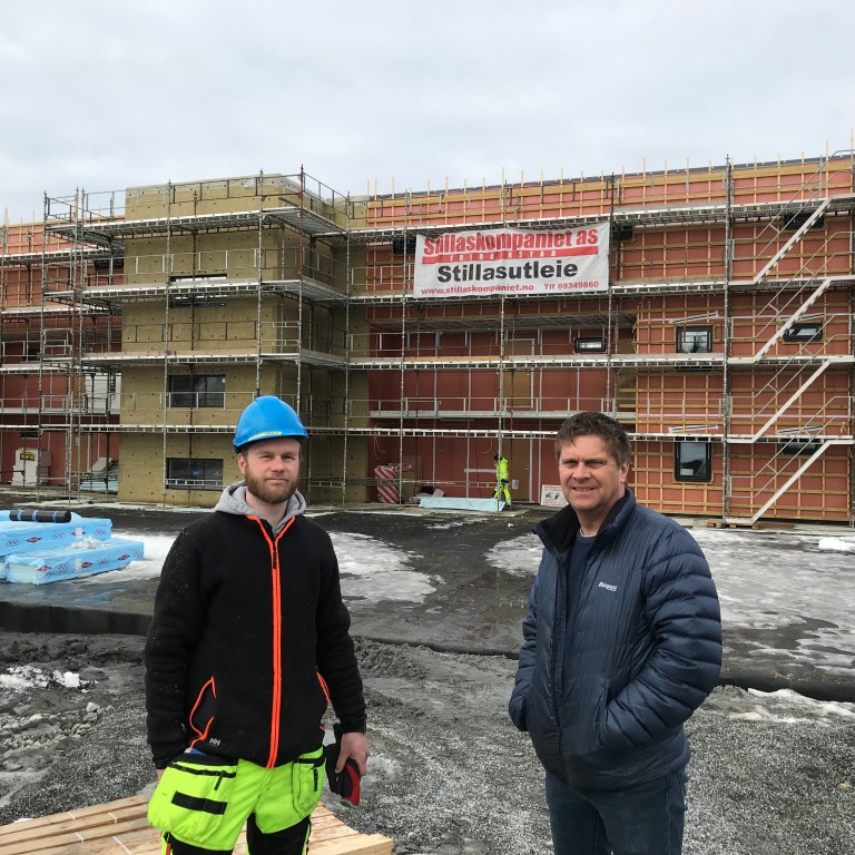 To håndverkere (én i blå hjelm og neongul arbeidshose) foran rødt stillas med snø.