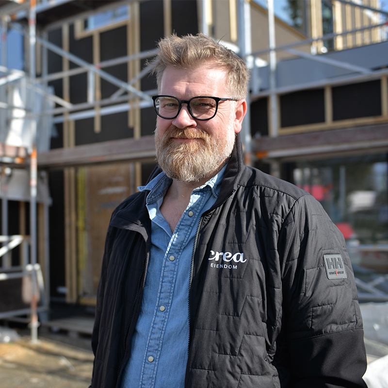Mann i svart jakke med “crea eiendom”-logo og blå skjorte foran byggeplass med stillas