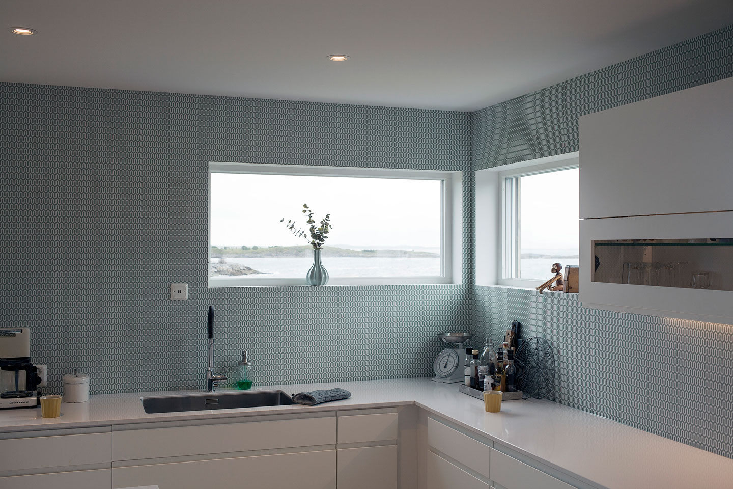 Lyst moderne kjøkken med hvit benkeplate, blågrå mønstret flisvegg, vindu mot sjøutsikt.