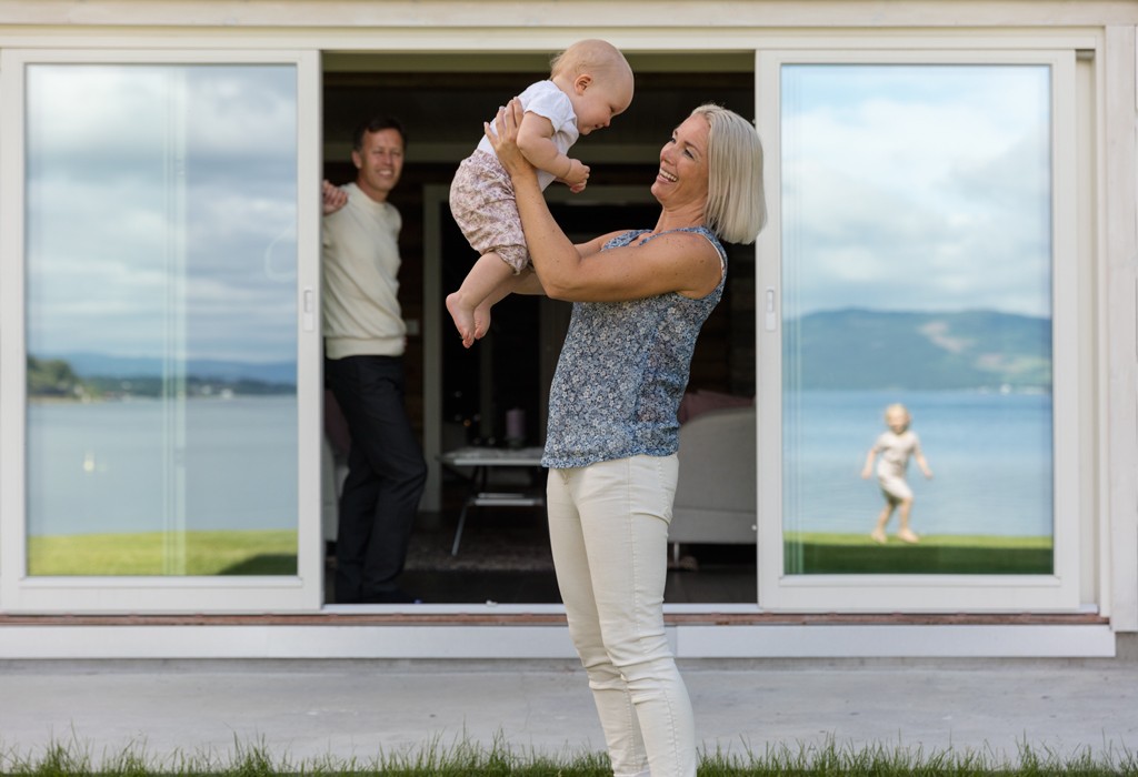 Mor i blå mønstret topp og hvite bukser løfter baby foran åpne skyvedører mot innsjø, barn leker.