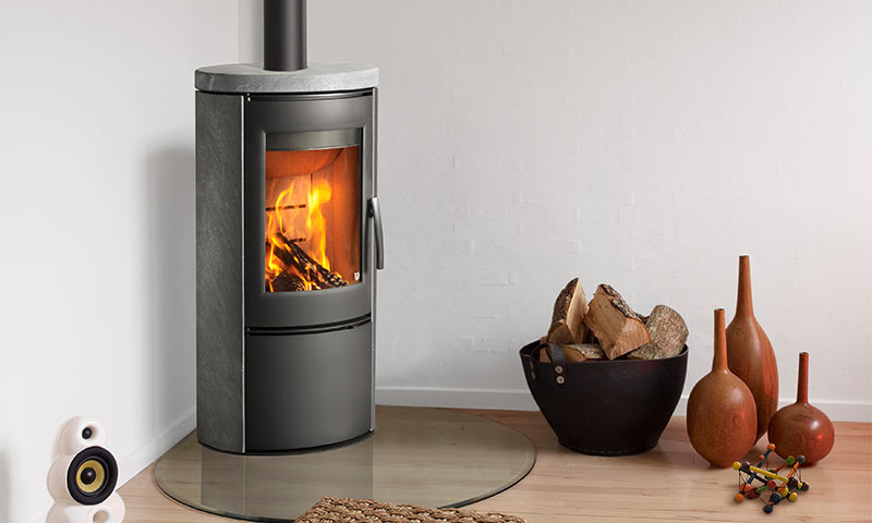 Sotgrå vedovn med ild i hjørne, brennved i svart kurv og tre oransje dekorvaser på lyst gulv