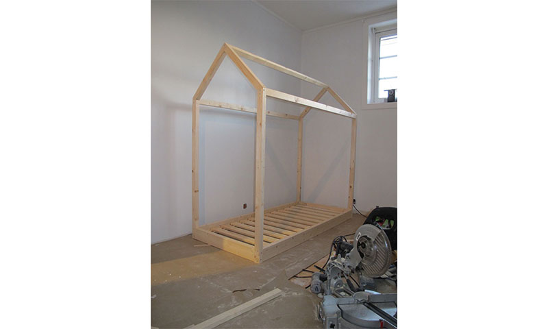 Uferdig furusengeramme formet som hus i hvitt rom, kappsag i forgrunnen.