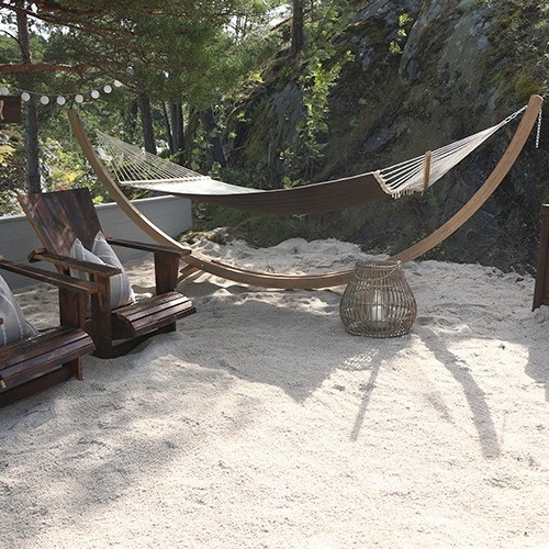 Beige hengekøye på lyst stativ over hvit sand ved to mørke trestoler og skog.