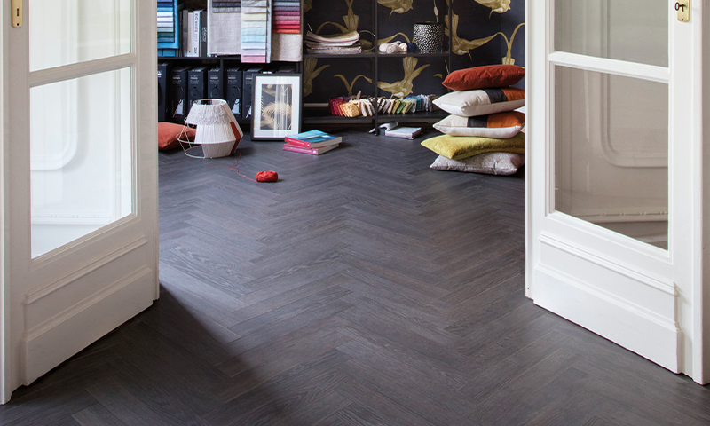 Åpne hvite dører til designstudio med mørk fiskebeinsparkett, fargerike stoffprøver og puter.