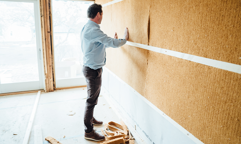 Mann i lyseblå skjorte monterer brun korkplate på vegg i uferdig, lyst oppussingsrom