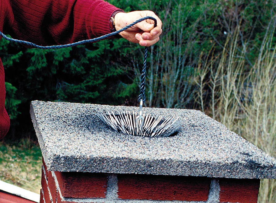 Hånd med blått tau holder rundt børstekost over grå pipelokk på rød teglstein mot grønn skog.
