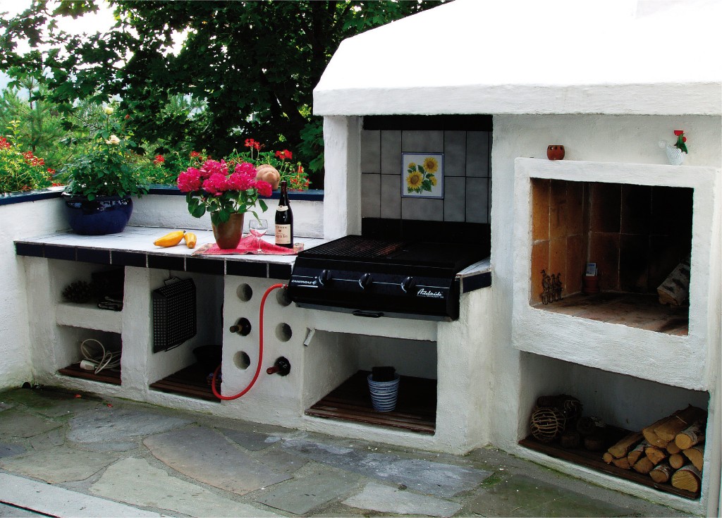 Hvit utendørs grillstasjon med svart gassgrill, rosa blomster, vinflaske, ved og grønt.