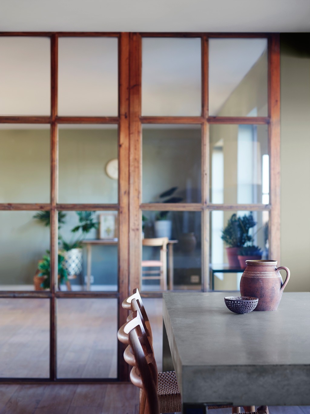 Grå betongbord med keramikkkanne og skål ved trestoler, foran glassdelt rom med grønne planter.