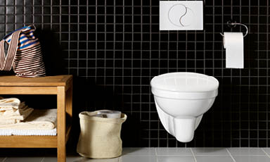 Sort flislagt baderom med hvitt vegghengt toalett, trebenk med håndklær, kurv og toalettrull.