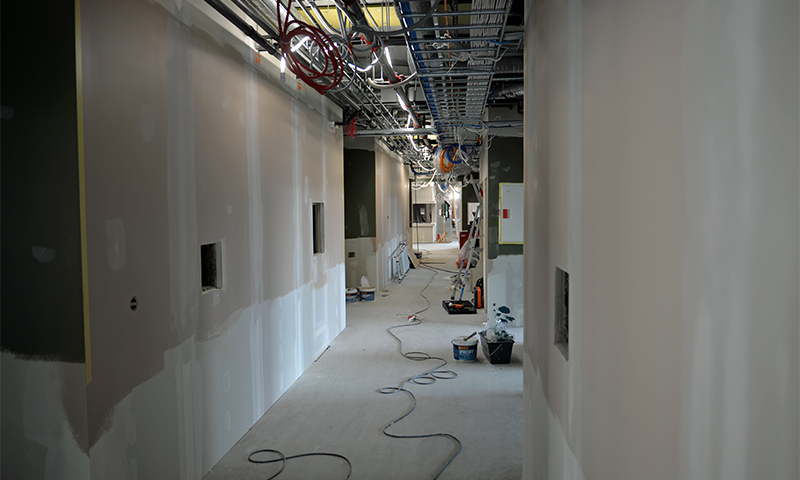 Lang korridor under oppussing med hvite gipsvegger, synlige takk abler og verktøy på gulvet.