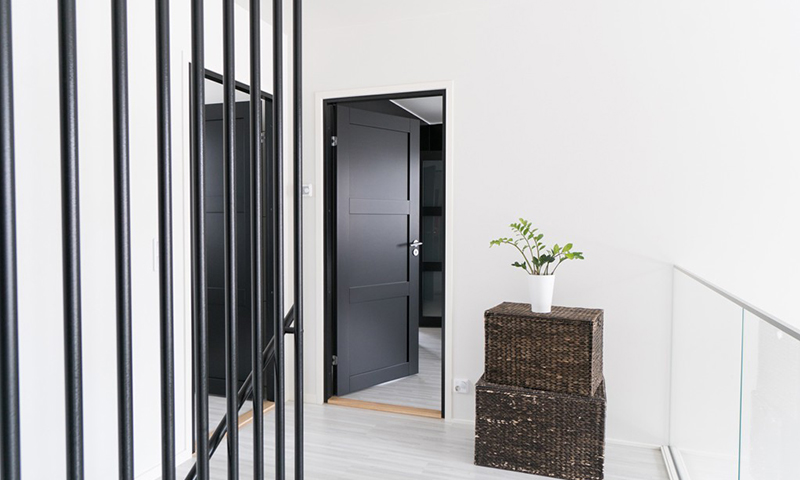 Lyst hvitt gangrom med svarte metallrekkverk, åpen mørk grå dør og kurv med grønn plante.