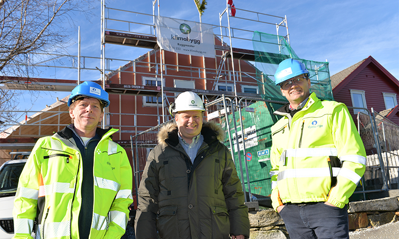 Tre byggearbeidere i gule jakker og hjelmer foran stillasert hus og blå himmel.