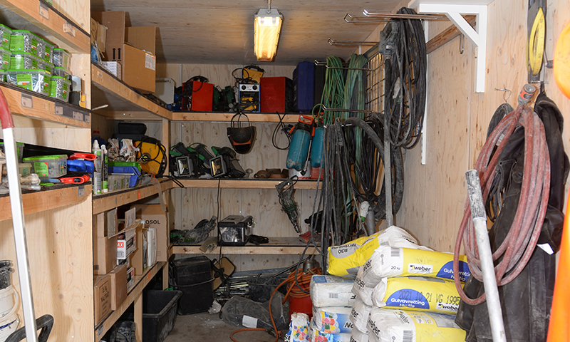 Lyst trebod med hyller fylt av verktøy, grønne kabler, svarte slanger og gule Weber-poser