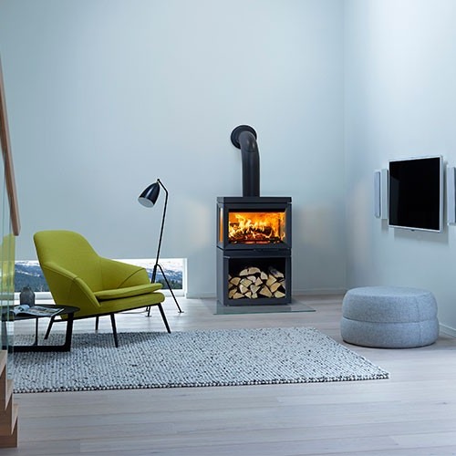 Moderne stue med lys grå vegger, vedovn med glødende ild, vedstabel, grønn lenestol, gulvlampe.