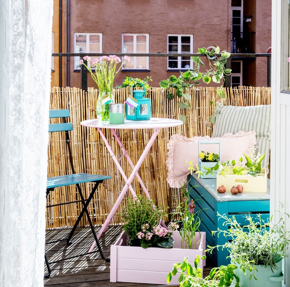 Lys balkong med rosa bord, blå stol, grønne planter, bambusgjerde og puter.