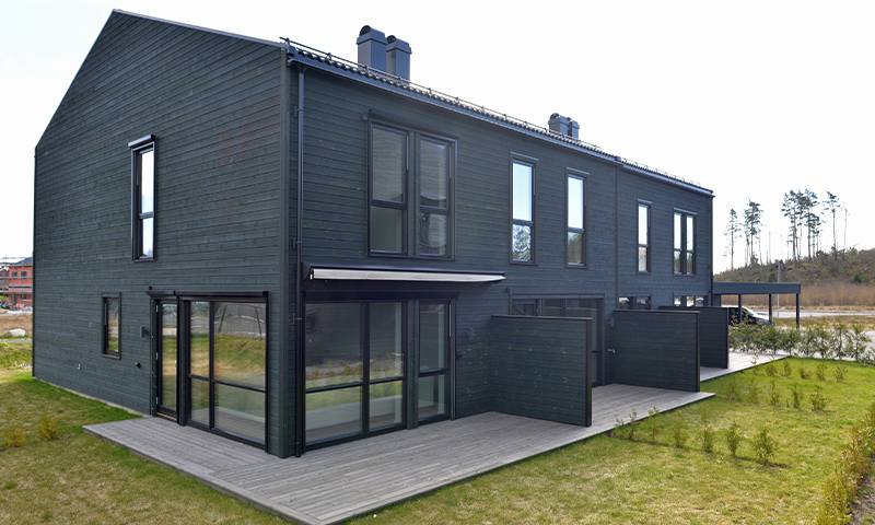 Svart toetasjes rekkehus i tre med store glassdører, terrasseplatting og gressplen.