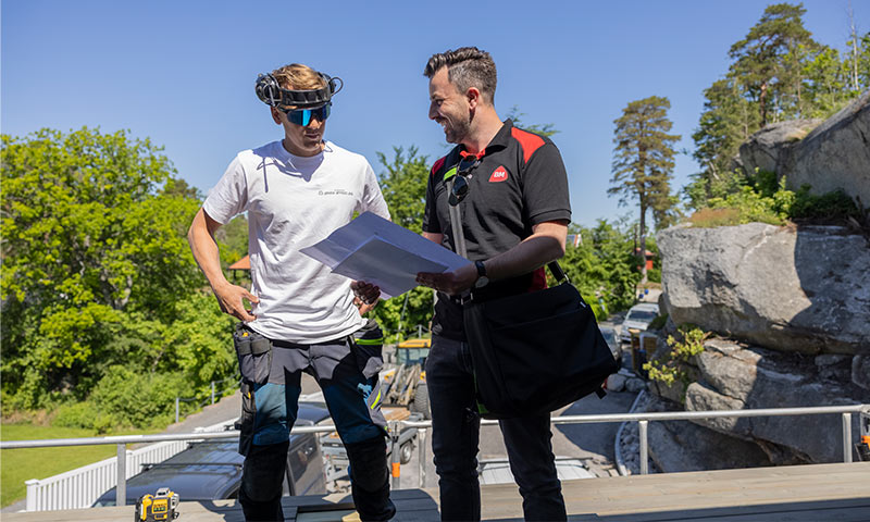 To håndverkere på tak under blå himmel, studerer papirer, med grønn skog og klippe bak dem.