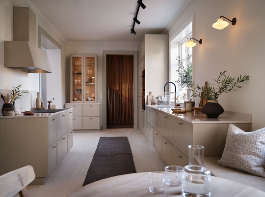 Moderne kjøkken med beige skap, mørk løper, vegglamper og grønne planter ved vinduet. Glasskrukker i forgrunnen.