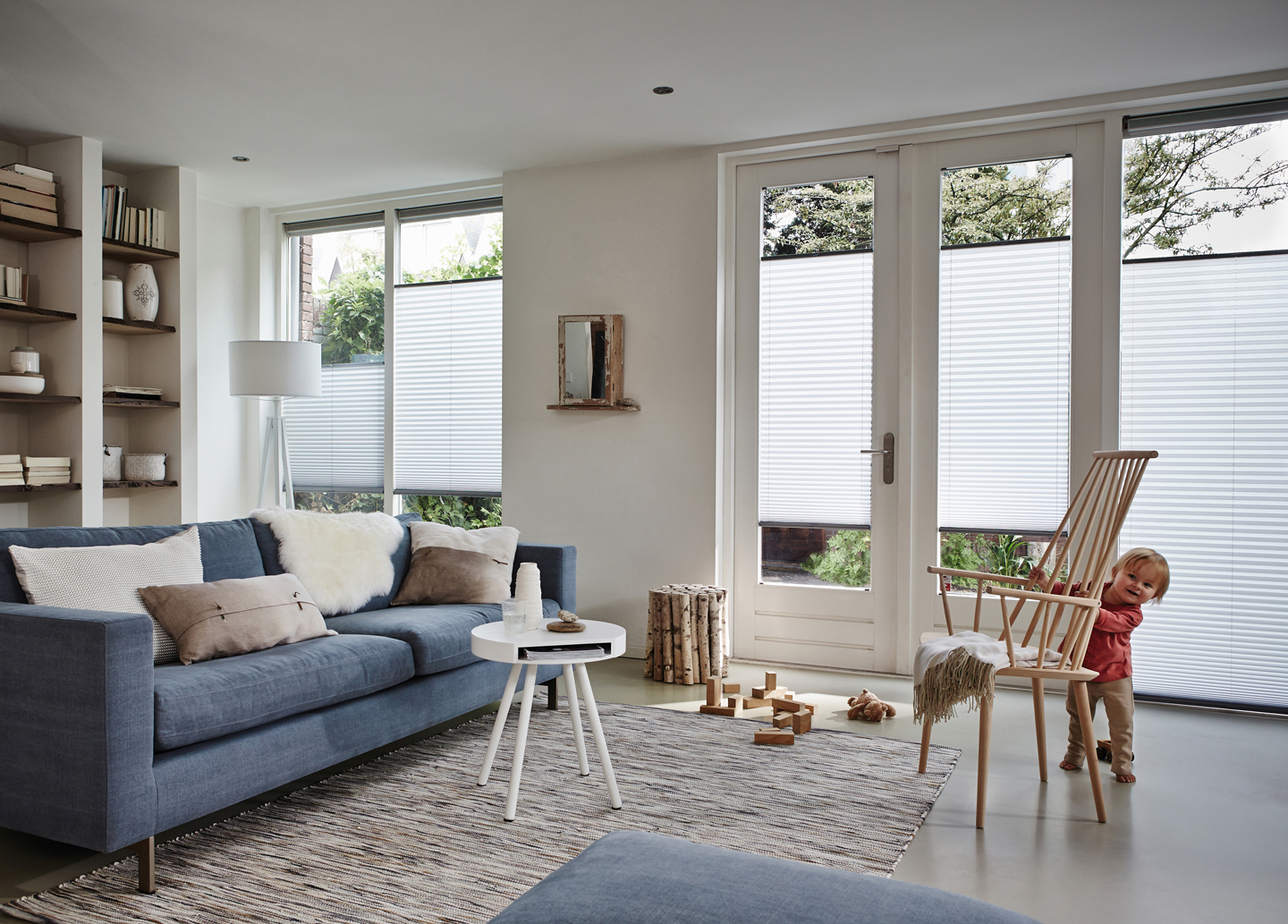 Lyse stue med blå sofa, hvitt rundt bord, trestol med pledd og hvite foldegardiner