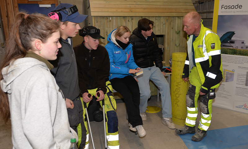 Håndverker i gul refleksjakke snakker med ungdommer i arbeidstøy foran trepanel og fasadebanner.