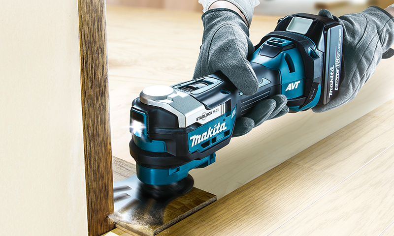 Arbeidshansker holder blå-svart Makita multiverktøy med LED-lys mot lyst trebord.