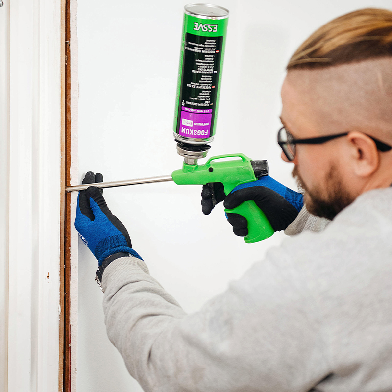 Person tetter dørkarm med grønn fugeskumspistol, blå hansker mot hvit vegg.