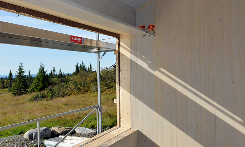 Åpent vindusfelt i uferdig trevegg, sollys på innside, utsikt til grønt landskap med furutrær.